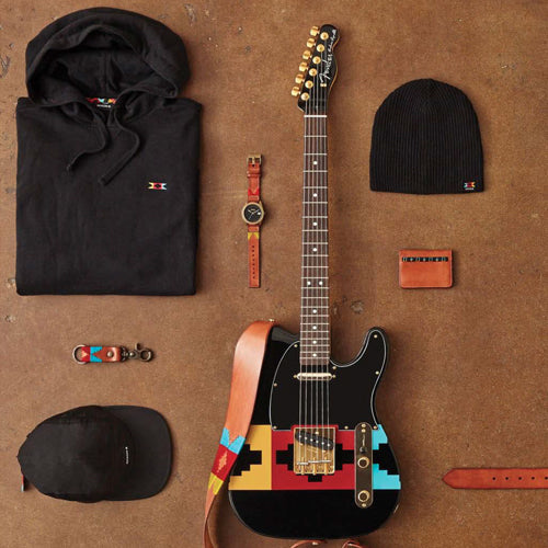 A black Fender Telecaster guitar with a colorful geometric strap is surrounded by a black hoodie, beanie, cap, leather wallet, watch, and keychain on a brown surface.