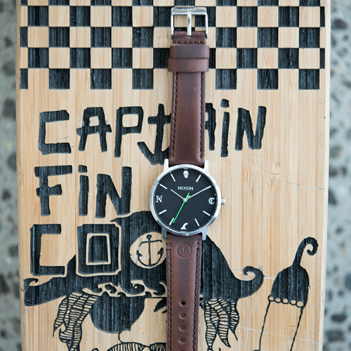 A Nixon analog watch sits on a decorated piece of wood with "Captain Fin" written on it.