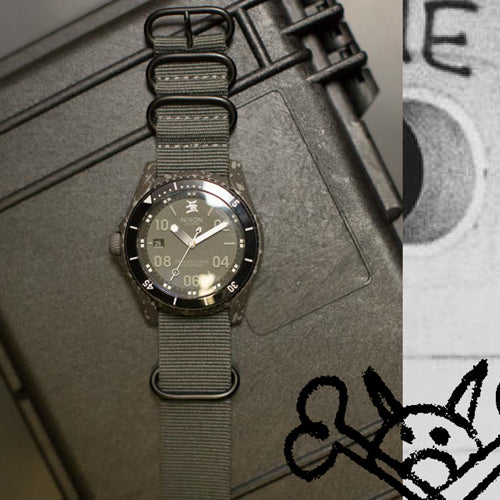 A dark grey Nixon watch with a fabric strap rests on a textured black case.