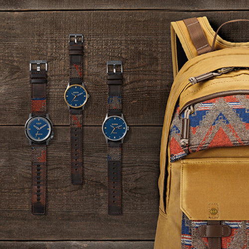 Three watches with blue faces and patterned leather straps hang on a wooden wall next to a tan backpack with a matching patterned pocket.