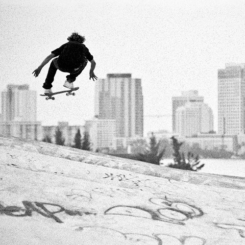 A black and white photo shows a skateboarder mid-air over a concrete surface with graffiti, with city buildings in the blurred background.