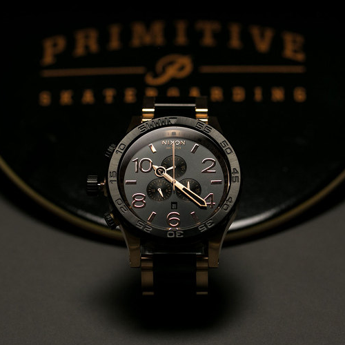 A black and gold Nixon chronograph watch with a dark grey face and rose gold accents sits in front of a blurred "Primitive Skateboarding" logo.