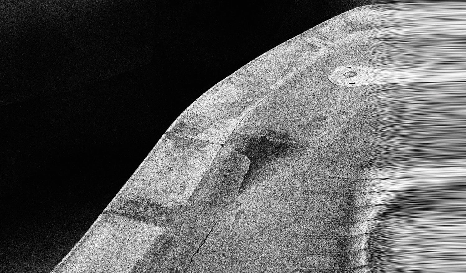 A grainy, black and white abstract image shows a curved concrete or stone structure with a circular element against a dark background.
