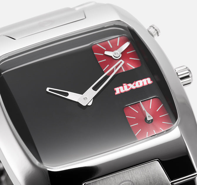 A close-up of a Nixon watch with a black face, silver hands, and two small red square dials.