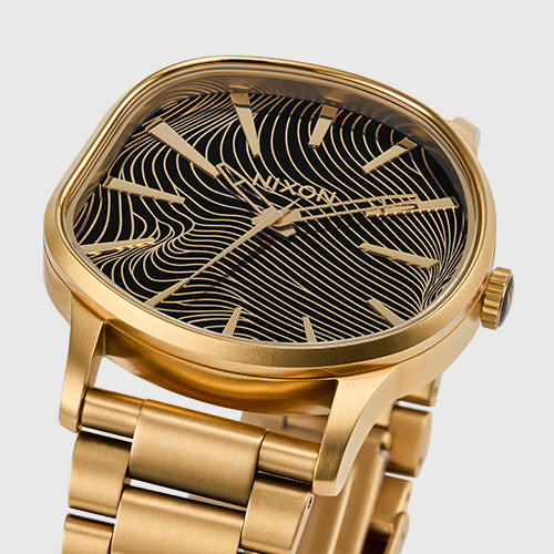 A gold Nixon watch with a black face featuring a wavy gold line pattern.