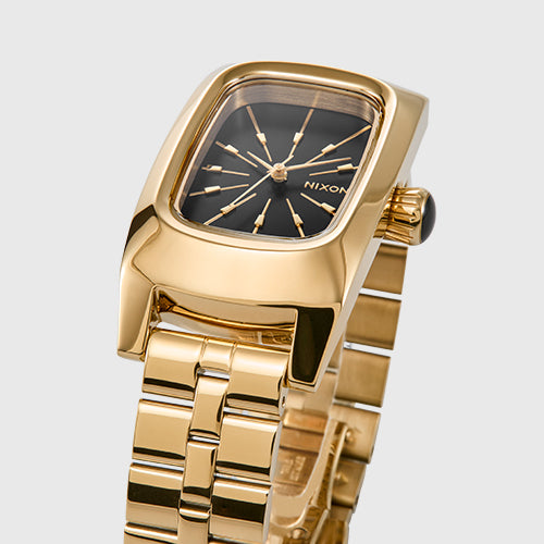 A close-up shot of a gold Nixon watch with a black rectangular face and gold hour markers.