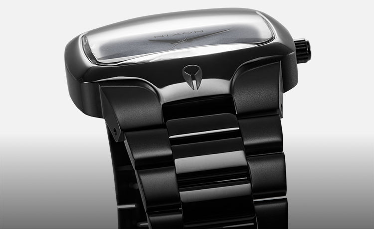 A close-up, angled shot of a sleek, all-black Nixon watch with a rectangular face and metallic band.