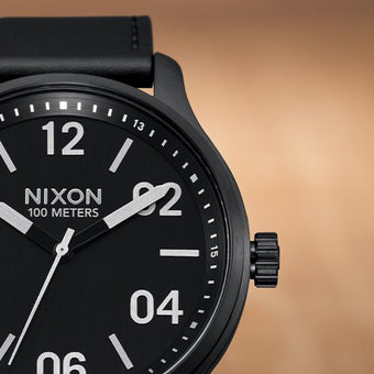 A close-up shot of a black Nixon analog watch with a black leather strap and white numerals.