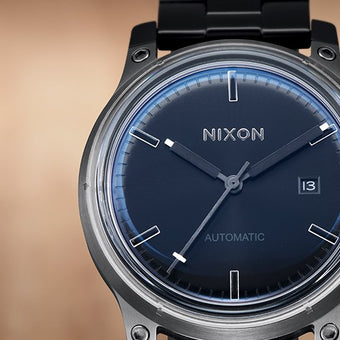 A close-up shot of a Nixon automatic watch with a dark blue face, silver hands, and a date display.