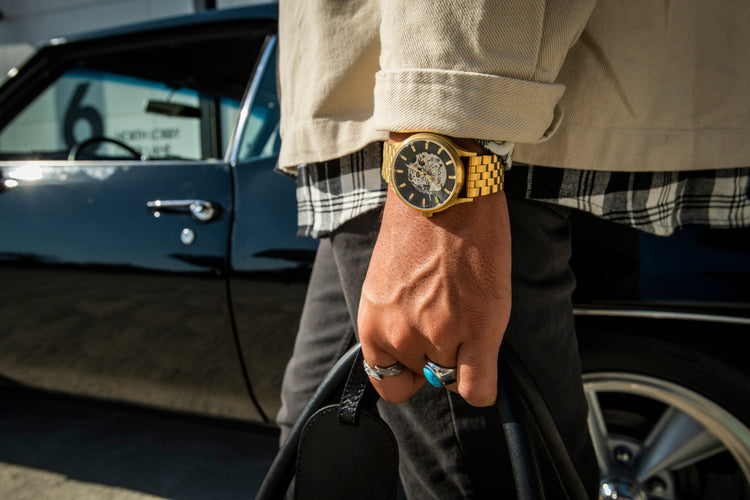Close-up of a person wearing a gold skeleton watch, plaid shirt, and beige jacket, holding a black bag next to a classic black car.
