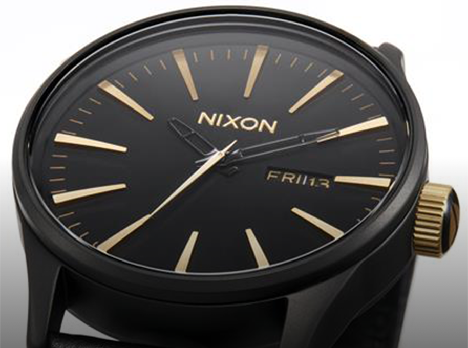 A close-up shot of a black Nixon watch with gold hour markers, hands, and crown, displaying "FRI 13" on the date window.